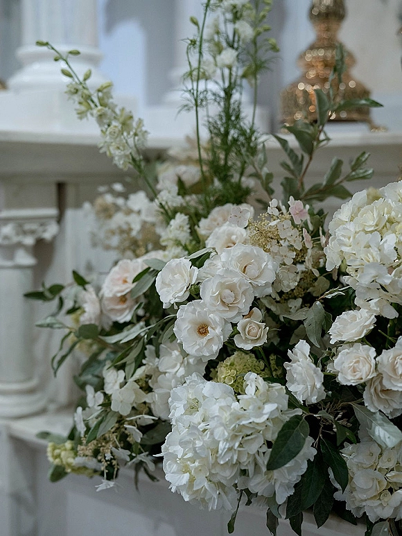 Wedding floral arrangement of white wedding flowers with roses and hydrangeas in a gold urn atop a white mantel against paneled wall