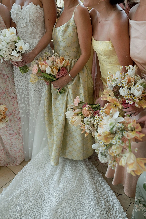 Bridesmaid bouquets of pastel tulips, daisies, and orchids held by mismatched dresses beside a lace wedding dress train in window light