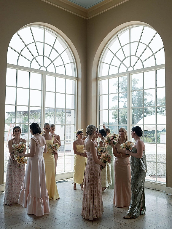 Bridesmaids portrait of friends holding bouquets and raising champagne flutes, in pastel dresses by arched windows with natural light