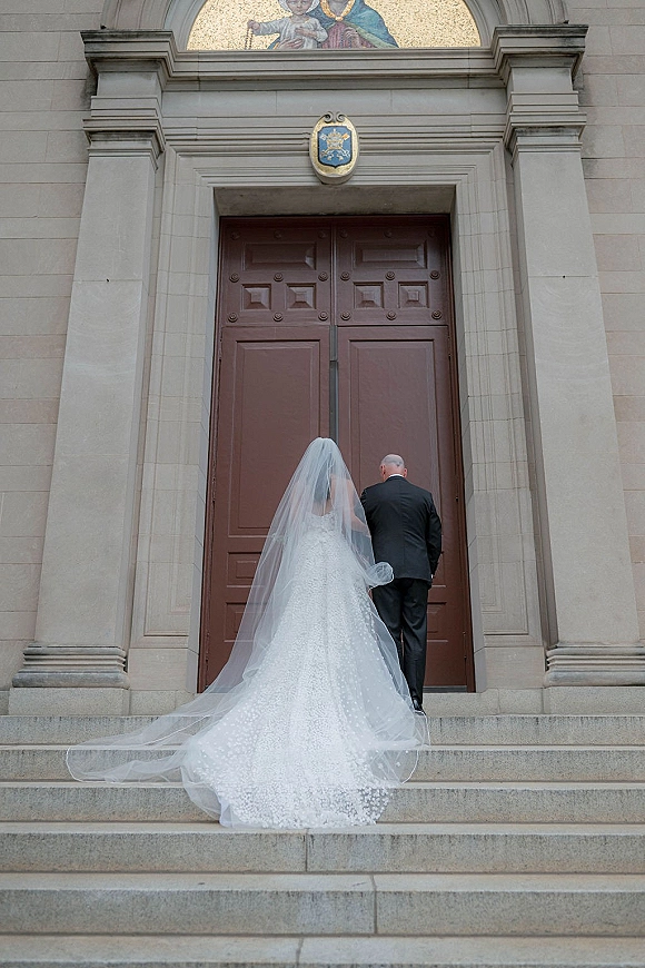 Wedding portrait of bride and groom from behind walking up steps, her long veil and dress train flowing toward grand wooden church doors