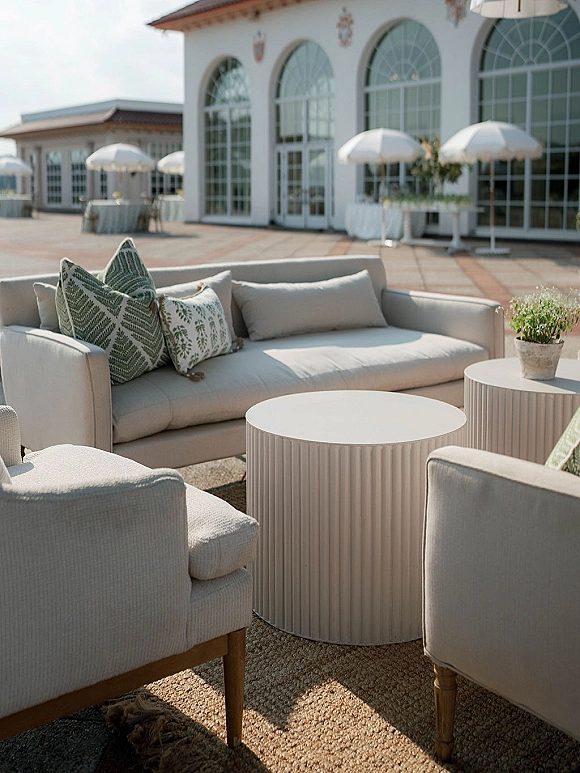 Wedding lounge seating with a cream sofa and upholstered chairs around a round coffee table on a stone patio beneath umbrellas and arched windows