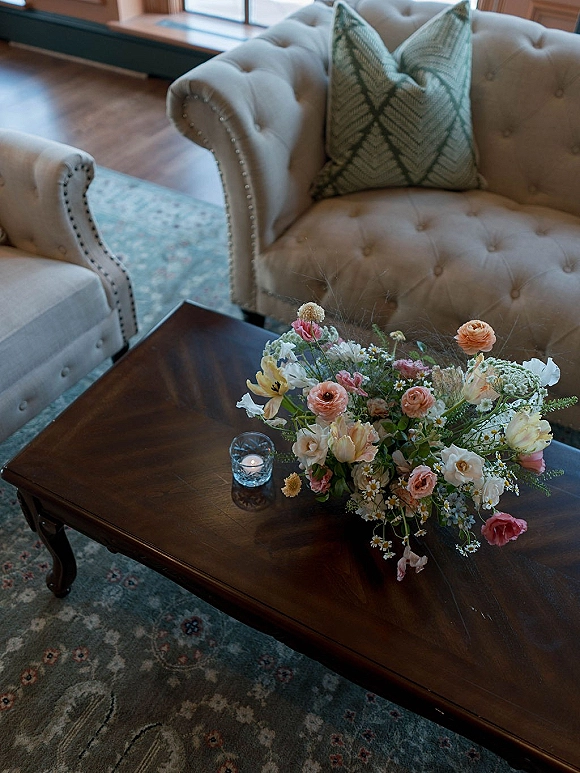 Wedding bouquet in garden style with blush and peach blooms on a wooden coffee table beside a glass votive candle in window light