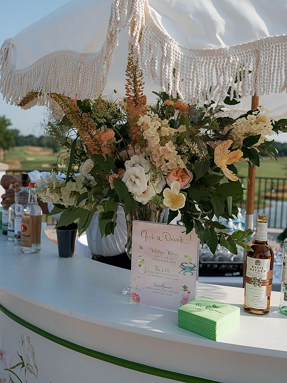 Wedding bar setup with a signature cocktail menu sign, liquor bottles, and lush floral arrangement under a fringe umbrella by a railing outdoors