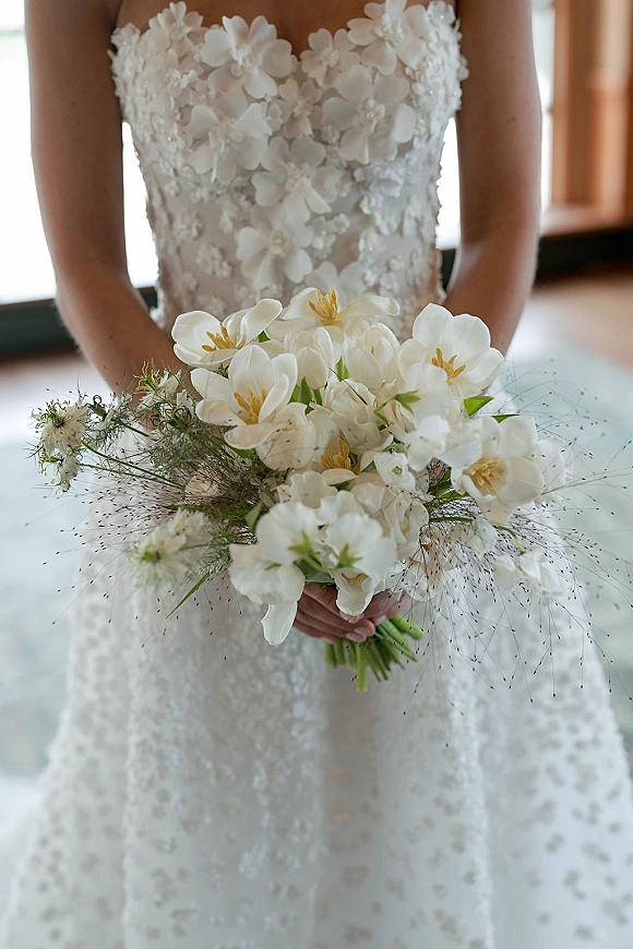 Bridal bouquet of white orchids with green stems held against a strapless lace wedding dress in soft indoor window light