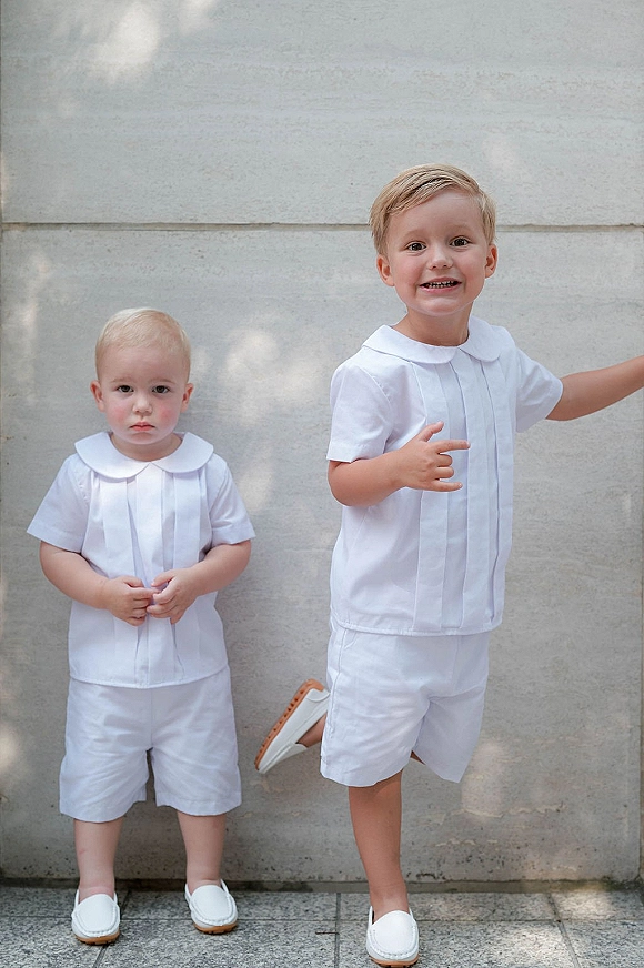 Ring bearer outfits on two boys in matching white short sets with peter pan collars, standing by a stone wall in dappled light