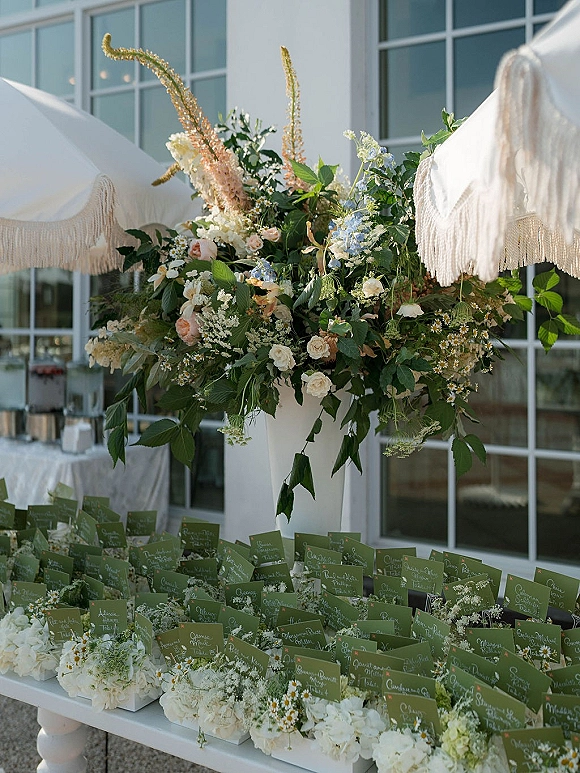 Wedding escort card display with calligraphy escort cards in a white planter, framed by pastel florals and baby’s breath on a patio by glass windows