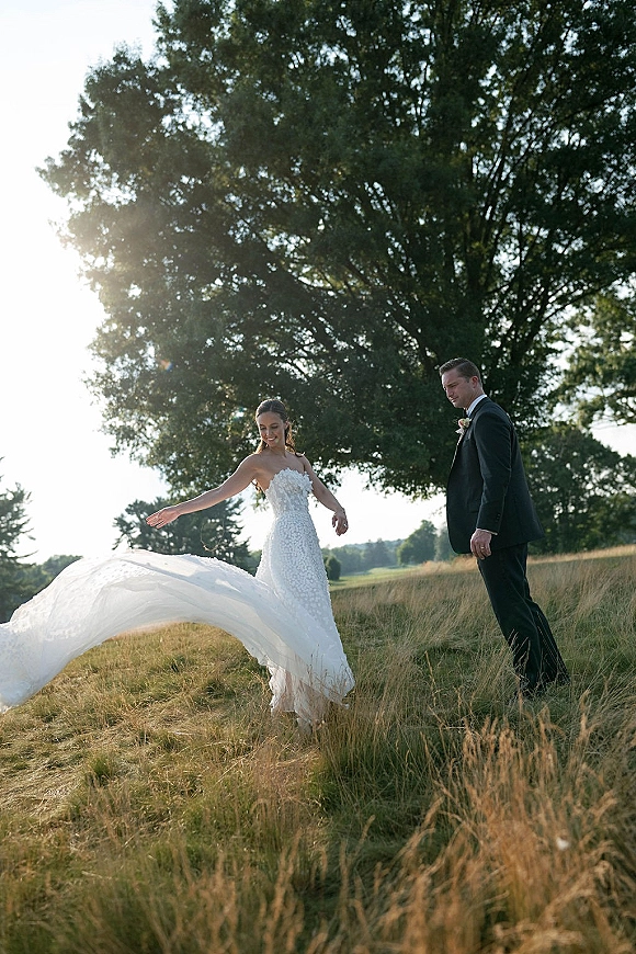 Couple portrait of bride twirling veil beside groom in black tuxedo, her strapless lace dress backlit in sunny meadow field