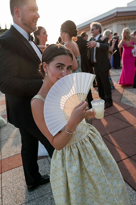 Wedding guest portrait of a couple in a black tuxedo and floral dress, holding a white folding fan and drink on a sunset terrace
