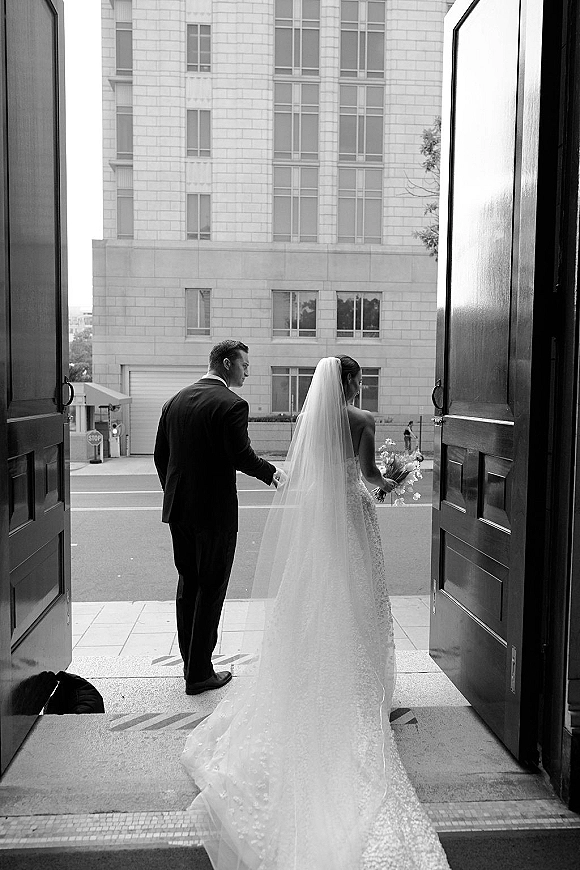 Wedding couple portrait in a black and white wedding photo, bride and groom holding hands from behind exiting open doors to a city street