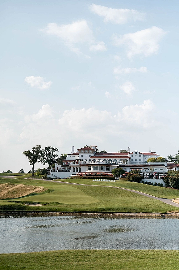 Outdoor ceremony setup with white ceremony chairs arranged on a lawn beside a pond at a golf course, clubhouse and clouds behind