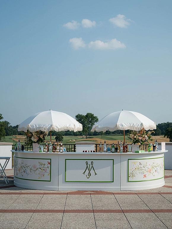 Outdoor wedding bar with a wedding cocktail bar setup under white patio umbrellas, floral panels and bottles on a terrace overlooking a golf course