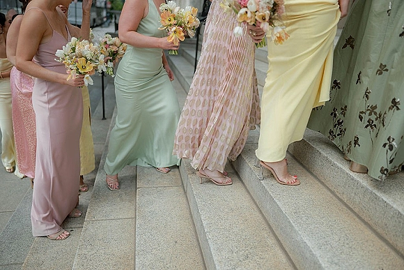 Bridesmaid dresses in pastel bridesmaid dresses, with bouquets and strappy heels, lined on stone steps along an outdoor walkway