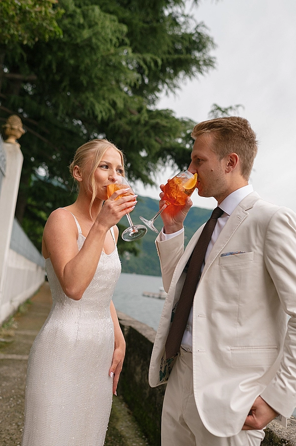 Couple portrait of bride and groom toasting with orange cocktails, bride in fitted dress and groom in beige suit by a mountain lake