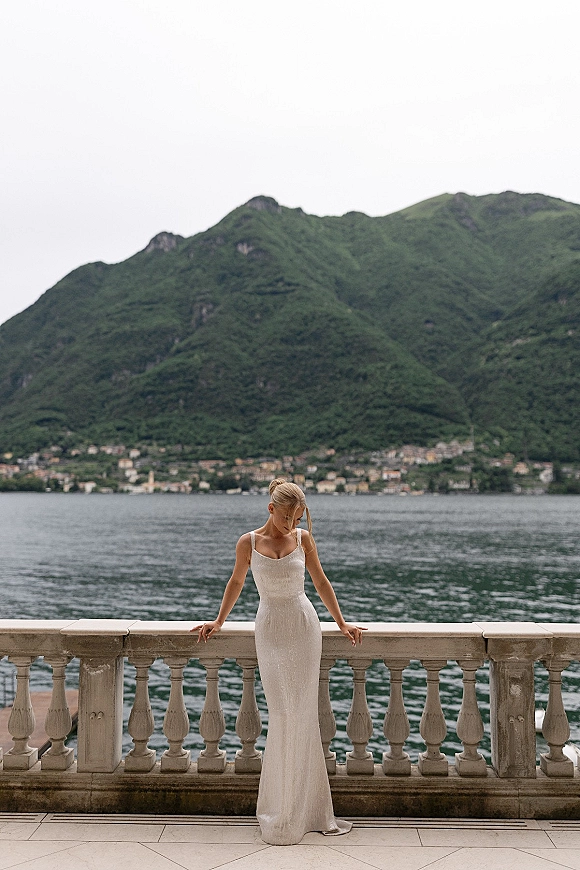 Bridal portrait of a bride leaning on railing in a fitted spaghetti-strap gown, looking down by a lakeside terrace with mountain backdrop