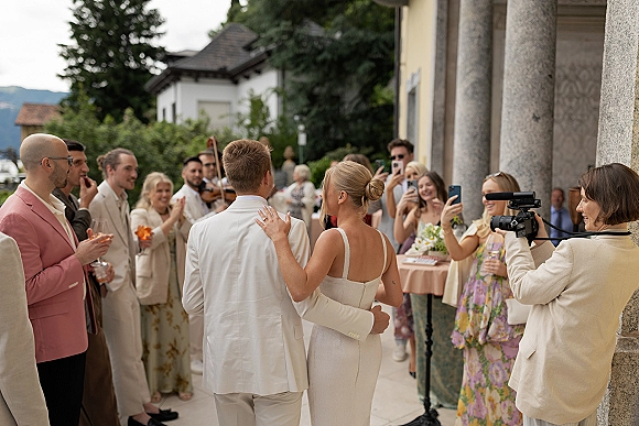 Wedding exit as newlyweds walking away arm in arm past cheering guests with phones raised, stone column venue entrance behind them
