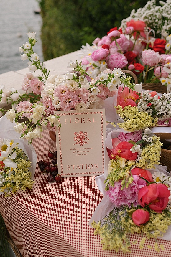 Wedding floral station with bouquets of roses, peonies, and daisies in wicker baskets on a gingham tablecloth, by water greenery backdrop