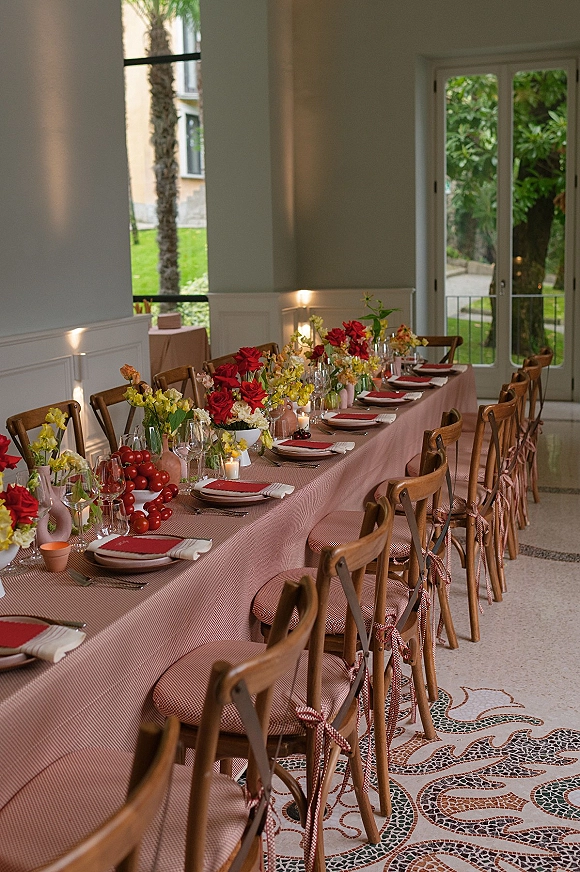 Reception tablescape with blush tablecloth on a long banquet table, red napkins, candles, and colorful florals in a bright room by garden-view windows