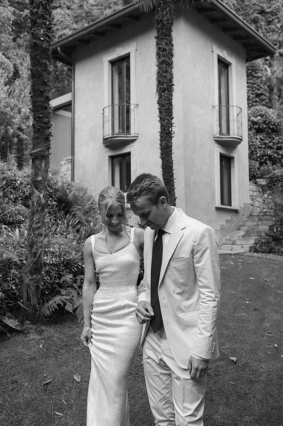 Couple portrait in a black and white wedding portrait style, bride in fitted dress and groom in light suit walking by palm trees and villa steps