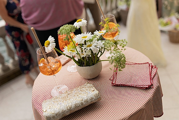 Cocktail table decor with a daisy centerpiece and aperol spritz wedding drinks on a red gingham cloth, guests blurred in reception behind