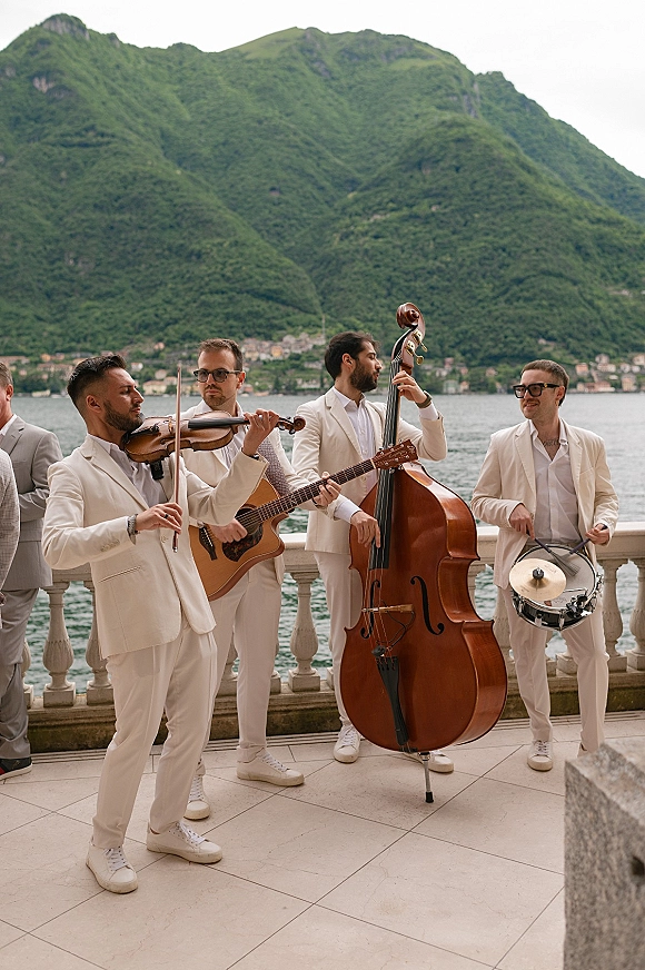 Wedding band in white suits playing violin, guitar, bass, and snare on a lakefront terrace with mountains under cloudy skies