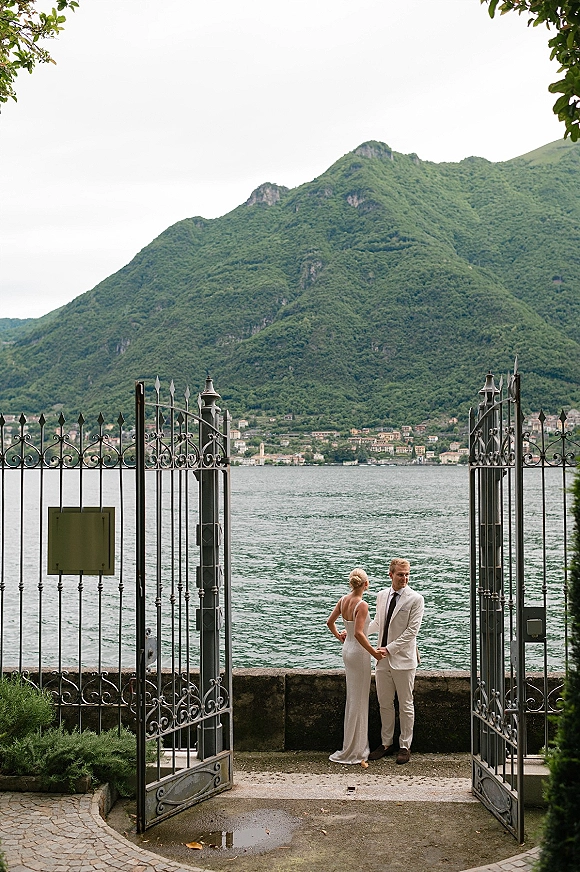 Couple portrait of bride and groom holding hands beside a wrought iron gate, with lake and mountains behind under an overcast sky