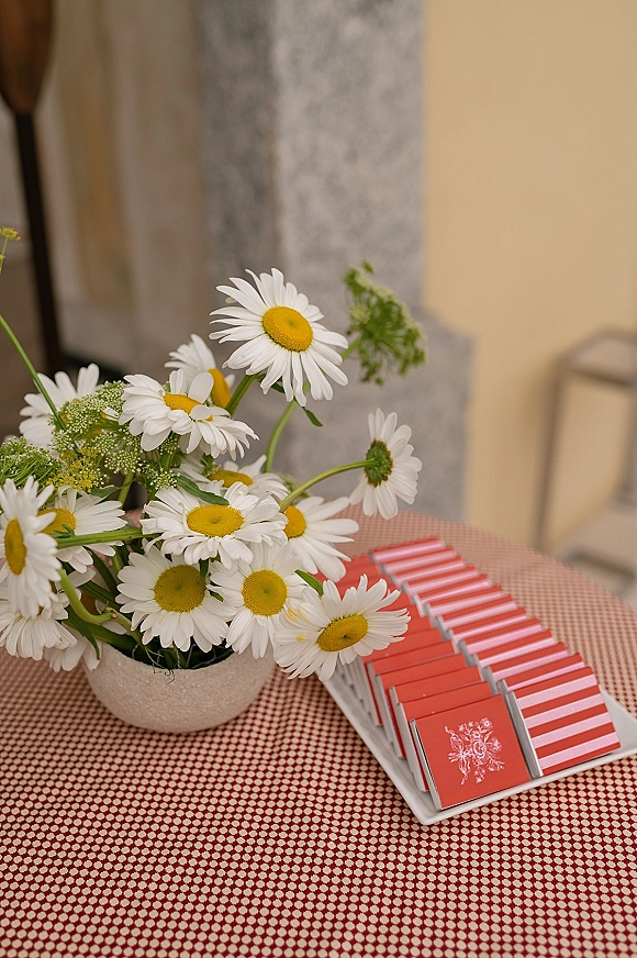 Wedding favors in striped wedding favor boxes arranged on a tray with daisy vase accent atop a red gingham tablecloth by a stone column