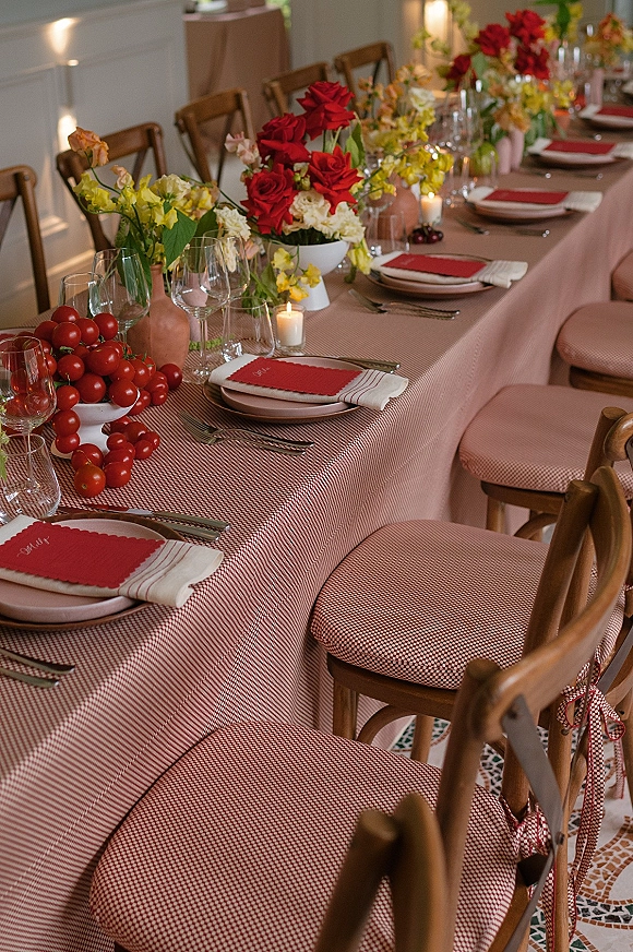 Reception tablescape with wedding head table decor featuring red floral centerpieces, candles, checkered cloth, red napkins, and cherries indoors
