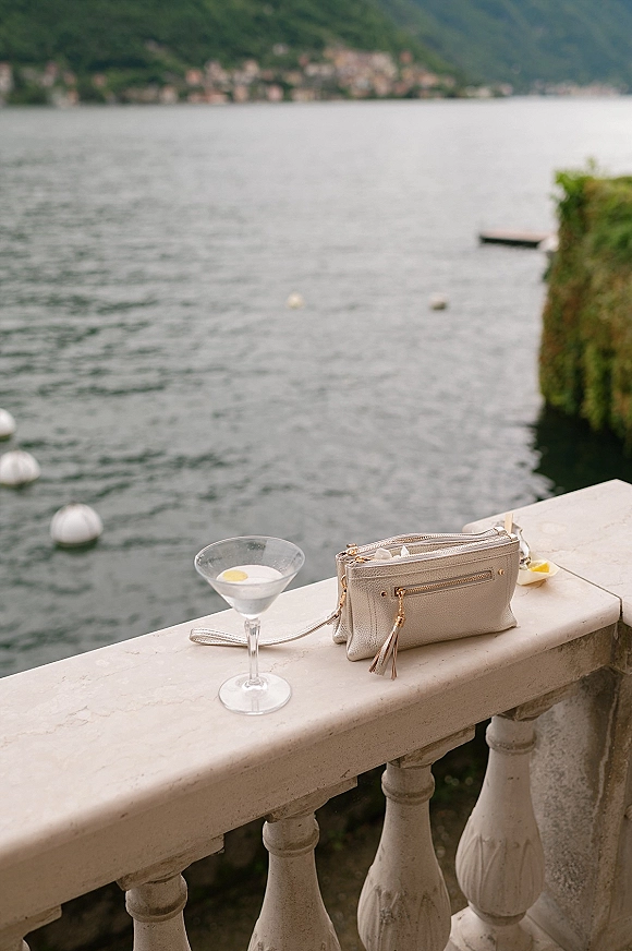 Wedding detail flatlay with a bridal clutch purse beside a cocktail glass with lemon slice on a stone balcony overlooking a lake and mountains