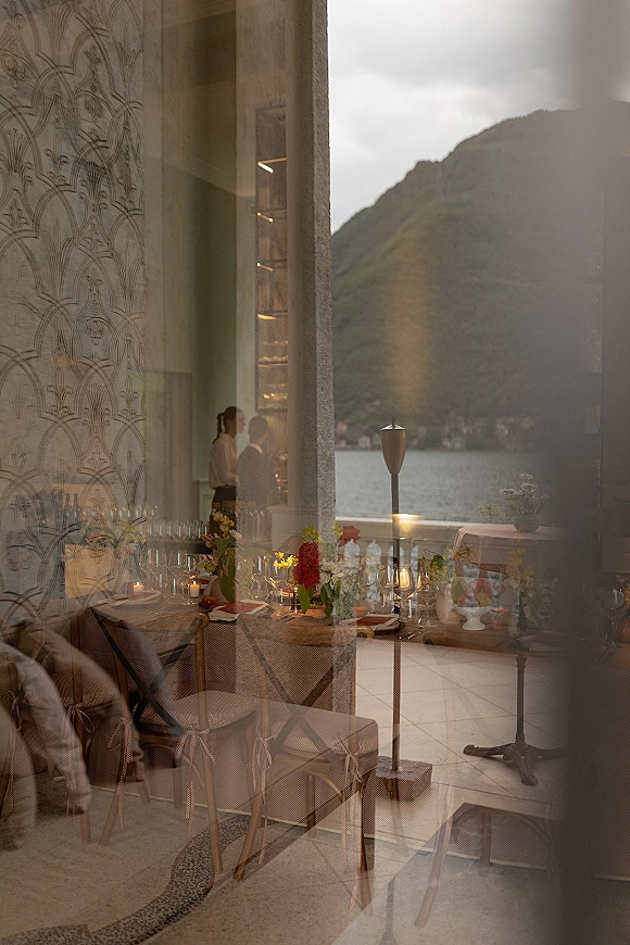 Reception tablescape with wedding reception table setting, floral centerpieces, candles and fruit on a patio beside arched windows and lake mountains view