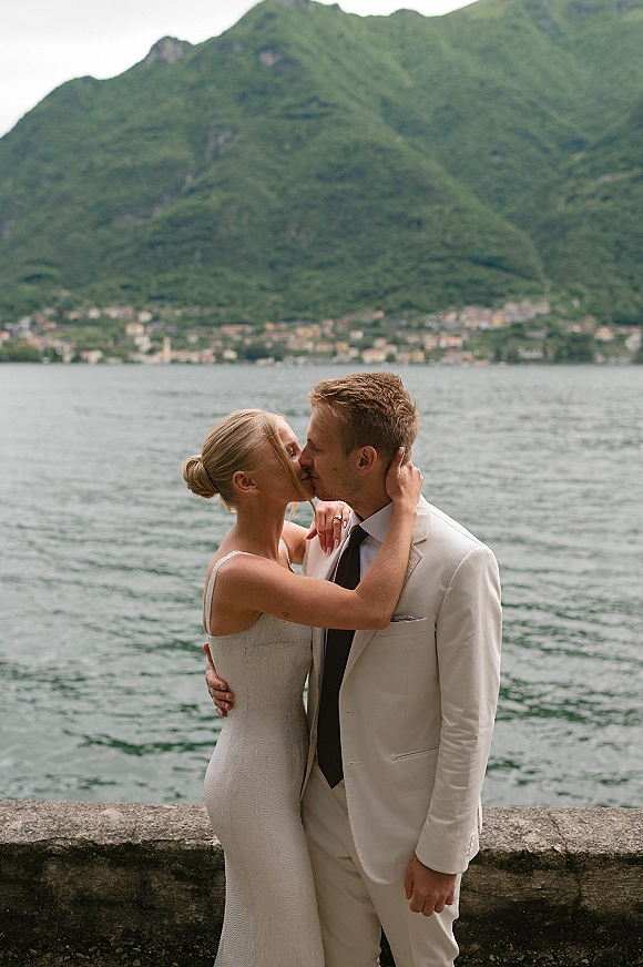 Wedding kiss portrait of bride and groom kissing in a sleeveless dress and light suit, with a mountain lake and stone wall behind them