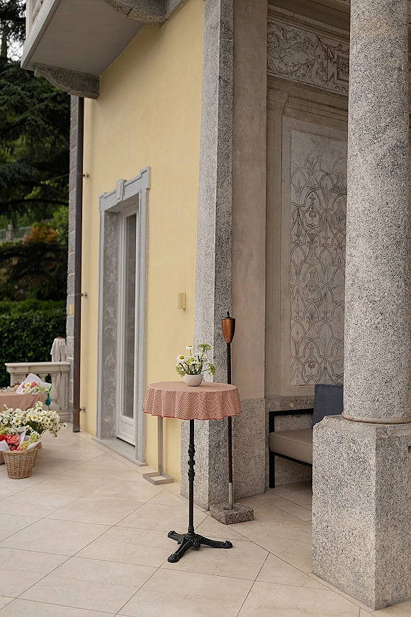 Wedding cocktail table with pink tablecloth, bud vase of daisies, and wicker basket of bouquets on a villa patio by stone columns