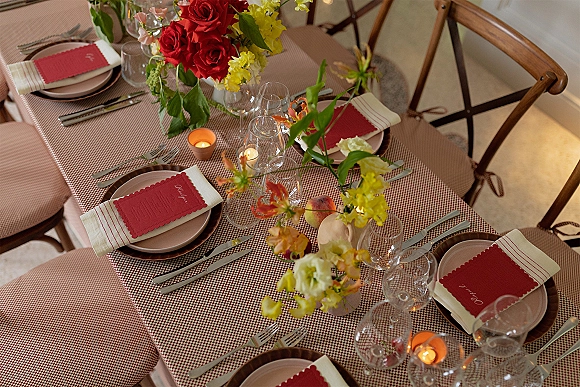 Reception tablescape with wedding table setting, floral centerpiece and bud vases, red menu cards on napkins, plates, glassware, votive candles on checked tablecloth