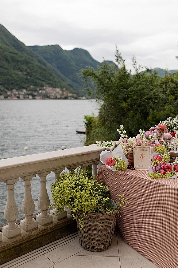 Wedding floral station with assorted arrangements and a flower bar wedding sign on a draped table, set on a stone terrace by a lake and mountains