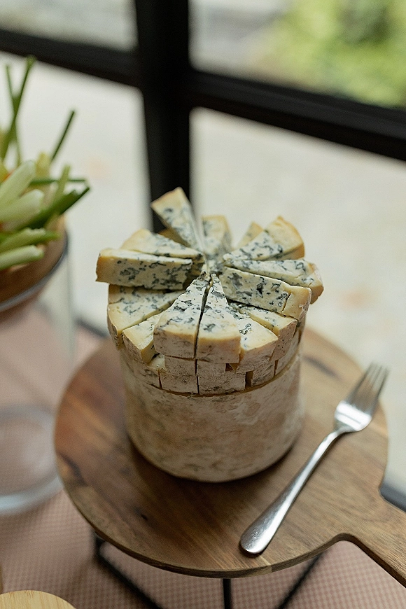 Cheese wheel appetizer with blue cheese wedges on a wooden serving board beside a vase of white flowers by window panes and greenery