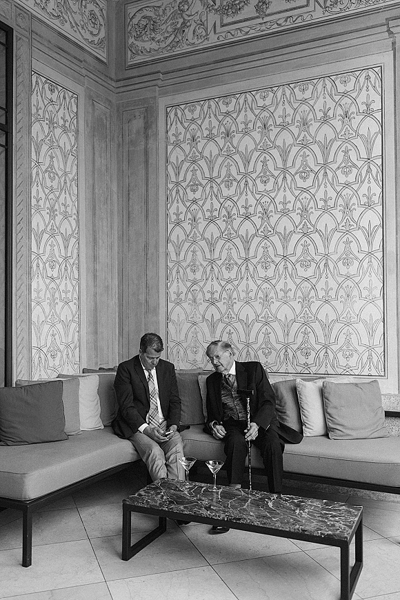Wedding guest moment as two men in suits chat in a lounge, one holding a cane, beside a sofa with martini glasses on a coffee table