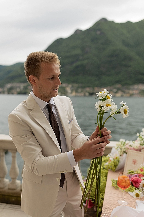 Groom portrait in a cream suit holding a daisy bouquet, standing on a stone terrace with lake and mountains under a cloudy sky
