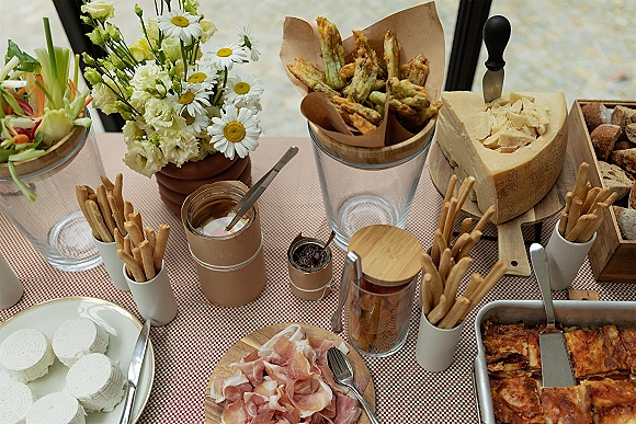 Wedding appetizer table with cheese wheel, breadsticks, prosciutto, rolls, and dipping sauces on red gingham tablecloth by a window light