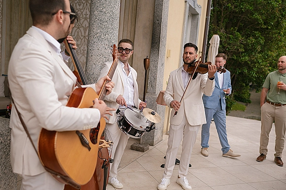 Wedding cocktail hour band in white suits playing acoustic guitar and upright bass on a patio terrace by stone columns and trees