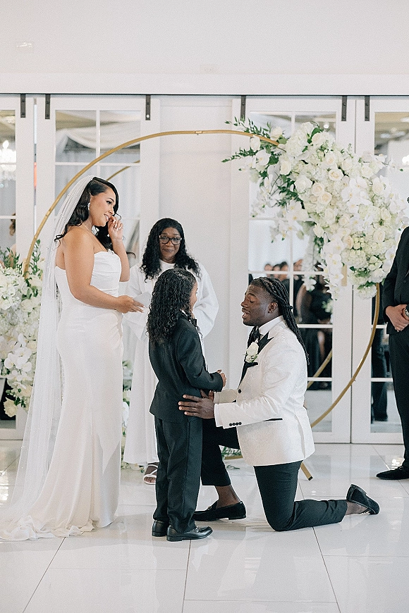 Ceremony moment as groom kneels before bride in strapless wedding dress and veil under a gold circular arch with white flowers indoors