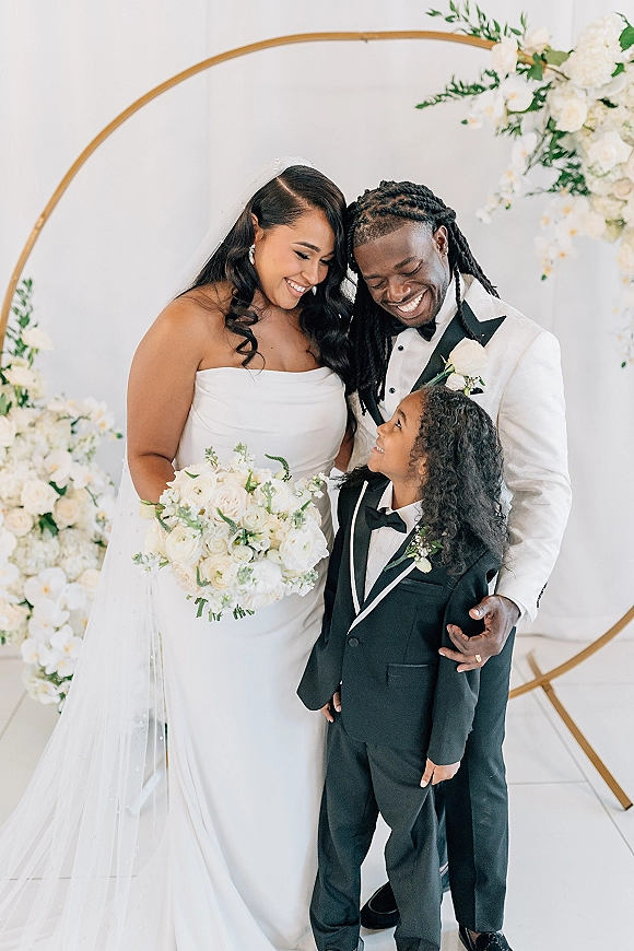 Family wedding portrait of bride and groom with child, bride holding a white bouquet under a gold circle arch against a white wall backdrop