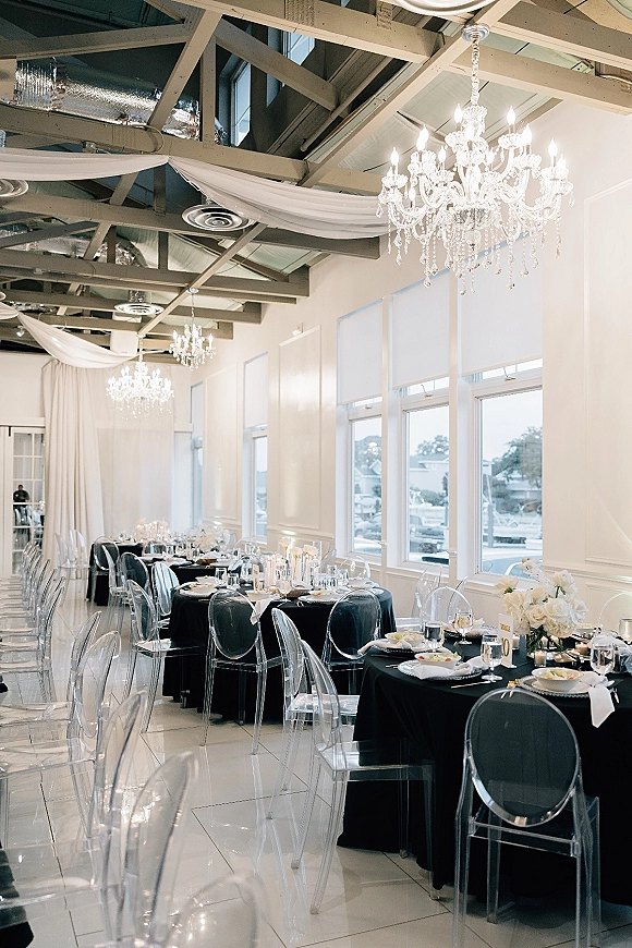 Reception tablescape with black and white reception decor, white floral centerpieces and candlelight beneath crystal chandeliers in a bright room