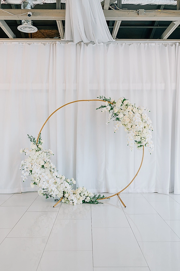Wedding ceremony backdrop with a circle wedding arch in gold, white drapery and asymmetrical white florals with greenery indoors