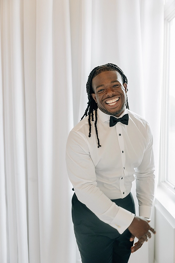 Groom portrait with a black bow tie, white dress shirt, and cufflinks, smiling in soft window light against white curtains and wall