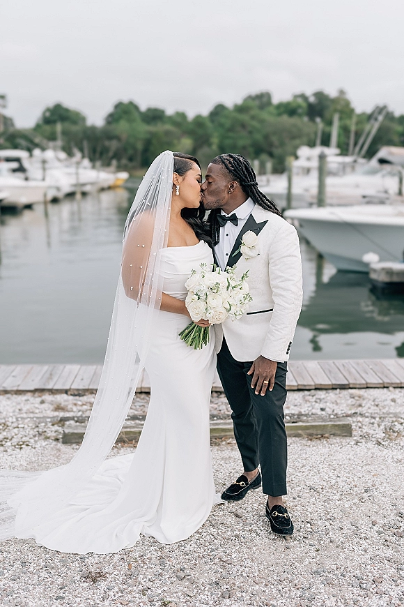 Wedding kiss portrait of bride and groom kissing at a marina dock, her long veil and white rose bouquet beside his white tuxedo jacket