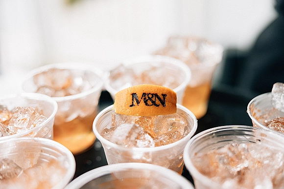 Wedding cocktail cups with amber drinks and ice, topped with citrus peel garnish, set on a dark tabletop near a blurred drink station