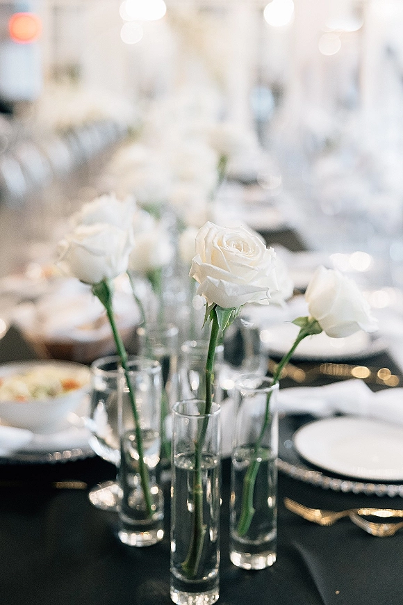 Reception tablescape with a black and white wedding table, white rose bud vases, glassware, and silver flatware under soft ambient lights