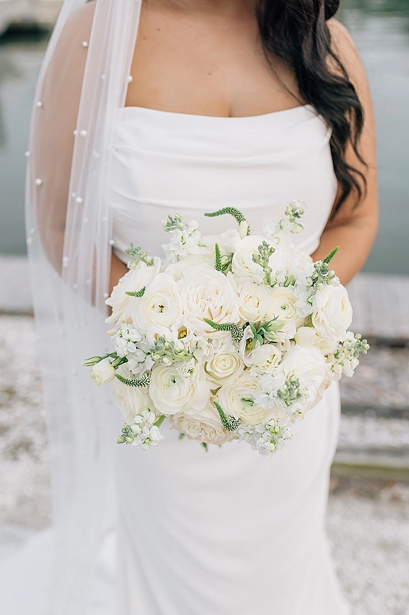 Bridal bouquet of white ranunculus and roses with greenery, held by a bride in a strapless dress and pearl veil on an outdoor walkway