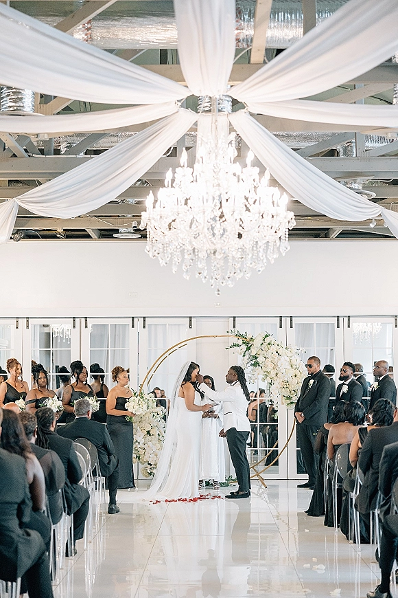 Wedding ceremony moment with couple exchanging vows under a circular white floral arch, long veil and rose petals lining the aisle indoors