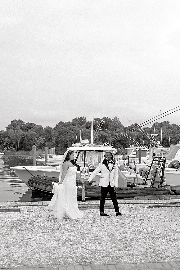 Couple portrait of bride in wedding dress and veil holding hands with groom in tuxedo jacket on a marina dock beside boats and water