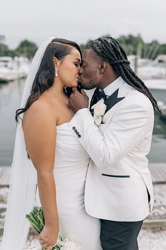 Wedding kiss portrait of bride and groom kissing, her veil and white rose bouquet visible, with boats at an overcast marina backdrop